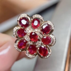 Puede incluir: Un anillo con forma de flor, con rub&iacute;es de talla ovalada y peque&ntilde;os diamantes. La montura del anillo es dorada, con cada rub&iacute; rodeado por un halo de diminutos diamantes. El dise&ntilde;o recuerda a una flor floreciente.