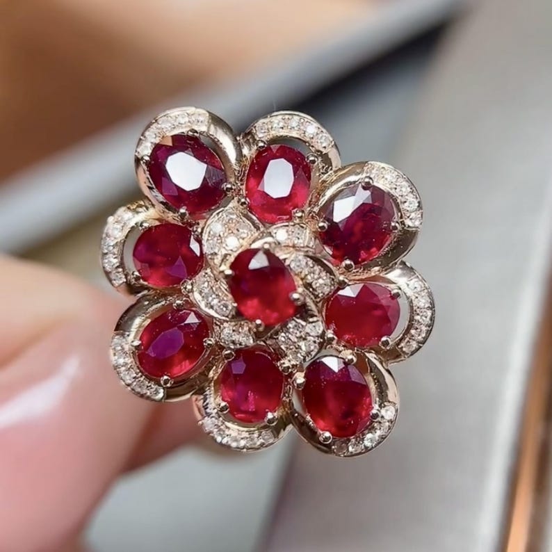 Puede incluir: Un anillo con forma de flor, con rub&iacute;es de talla ovalada y peque&ntilde;os diamantes. La montura del anillo es dorada, con cada rub&iacute; rodeado por un halo de diminutos diamantes. El dise&ntilde;o recuerda a una flor floreciente.