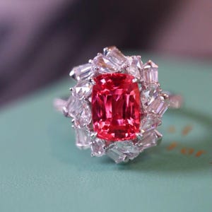 May include: A close-up of a ring featuring a large, rectangular, vibrant red gemstone surrounded by smaller, clear, rectangular and baguette-cut stones. The ring's band is silver-colored.