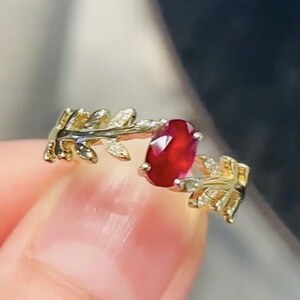 Può includere: Un anello d'oro con una pietra preziosa ovale rossa incastonata in un design a foglia. Le foglie sono accentate da piccole pietre preziose trasparenti.