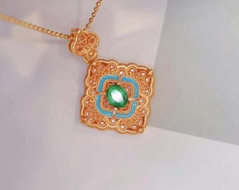 May include: Gold-toned pendant necklace featuring a square pendant with intricate filigree detailing. The pendant has a turquoise enamel design and a central oval green gemstone. The necklace chain is delicate and gold-colored.