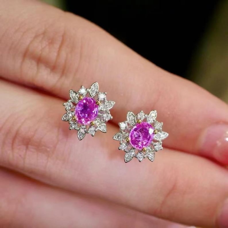 May include: A pair of pink sapphire and diamond earrings. Each earring features a pink oval sapphire surrounded by a halo of round diamonds set in a white gold setting.