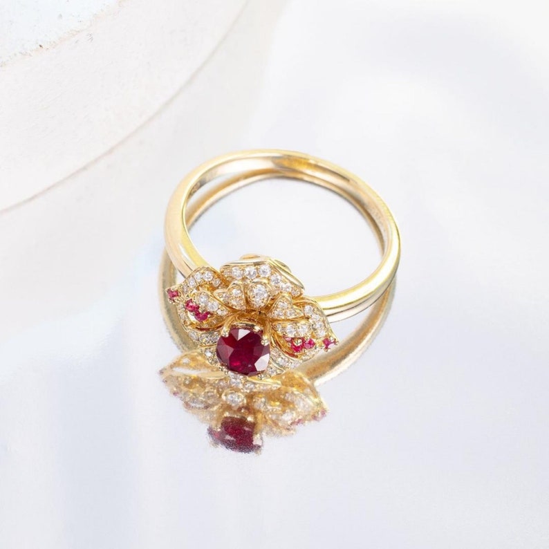 Puede incluir: Un anillo de oro con un dise&ntilde;o de flor de rub&iacute; y diamantes. El anillo est&aacute; colocado sobre una superficie blanca.