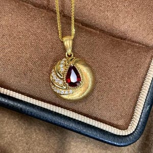 May include: A gold pendant necklace featuring a teardrop-shaped, deep red gemstone. The pendant is surrounded by small, sparkling white stones and a textured gold design. The necklace is displayed in a brown jewelry box.