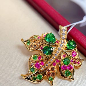 Puede incluir: Broche dorado con forma de hoja, adornado con piedras preciosas verdes, rosas y transparentes. El broche presenta un diseño detallado con múltiples piedras preciosas de varios tamaños y colores, engastadas en metal dorado.