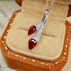 May include: A silver necklace with a pendant featuring two teardrop-shaped red gemstones and small clear gemstones. The pendant is in the shape of two stems.