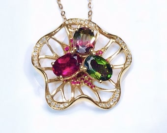 May include: A gold pendant necklace with a flower-shaped design. The pendant features three oval-shaped gemstones: a red, a green, and a multi-coloured stone. The flower design is outlined with small, clear stones.