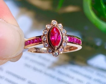 Op de afbeelding: Een ring van roségoud met een marquise-vormige robijn omringd door een halo van kleine diamanten. De band is bezet met een rij kleine robijnen.