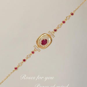 May include: Gold bracelet with a square-shaped pendant featuring a red gemstone surrounded by smaller clear gemstones. The bracelet has a chain with alternating red and clear gemstones.