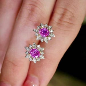 May include: A pair of pink sapphire and diamond earrings. Each earring features a pink oval sapphire surrounded by a halo of round diamonds set in a white gold flower design.