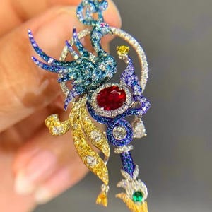 Puede incluir: Broche de joyería con forma de pájaro, adornado con piedras preciosas. Presenta una gran gema roja ovalada, rodeada de piedras azules, verdes, amarillas y blancas. El cuerpo del pájaro es una mezcla de piedras azules y verdes, con pico y cola dorados.