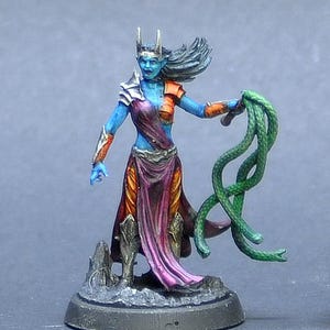 May include: A blue-skinned female miniature figure with long black hair, wearing a red and orange dress and holding a green snake. The figure is standing on a black base. A red and black 20-sided die is in the background.