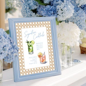May include: A light blue framed sign with the text "Signature Cocktails" and illustrations of a Mojito and Paloma. The frame has a woven pattern and is surrounded by blue and white flowers.