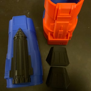 Puede incluir: Un conjunto de piezas impresas en 3D para un disfraz o un accesorio. Las piezas son azules, naranjas y negras. La pieza azul es una pieza larga y cil&iacute;ndrica con un detalle negro en el lateral. La pieza naranja es una pieza m&aacute;s corta y cil&iacute;ndrica con un dise&ntilde;o m&aacute;s complejo. Las piezas negras son dos piezas peque&ntilde;as y planas.