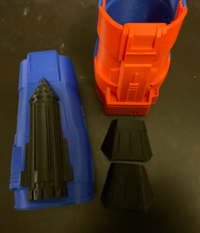 Puede incluir: Un conjunto de piezas impresas en 3D para un disfraz o un accesorio. Las piezas son azules, naranjas y negras. La pieza azul es una pieza larga y cil&iacute;ndrica con un detalle negro en el lateral. La pieza naranja es una pieza m&aacute;s corta y cil&iacute;ndrica con un dise&ntilde;o m&aacute;s complejo. Las piezas negras son dos piezas peque&ntilde;as y planas.