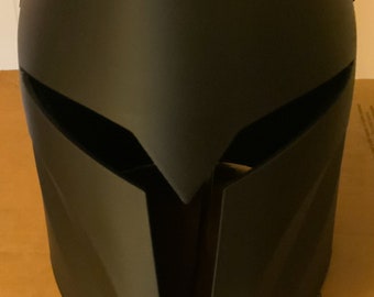Puede incluir: Un casco negro mate con un diseño angular distintivo. El casco presenta una visera puntiaguda y una parte superior lisa y curvada. La forma general es aerodinámica y futurista, lo que sugiere un tema de ciencia ficción o cosplay.