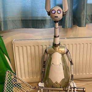 May include: A white and green robot with large ears and purple eyes holds a long, silver stick with a net attached to the end. The robot is standing on a brown floor in front of a white radiator.