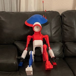May include: A red, white and blue robot with a blue dome-shaped head and black accents. The robot is sitting on a black leather sofa.