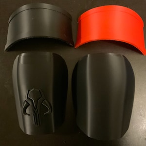 Puede incluir: Cuatro piezas impresas en 3D para un disfraz. Dos piezas negras tienen forma de espinilleras, una tiene un s&iacute;mbolo mandaloriano. Otras dos piezas son curvas y tienen forma de casco, una es negra y la otra es roja.