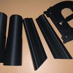 May include: Black plastic components, possibly for a mechanical device, arranged on a tan surface. The parts include two cylindrical pieces, two angled panels, and a curved, slotted piece. The items are all black.