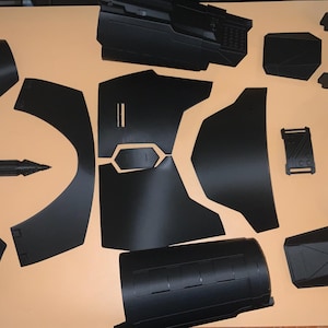 May include: A set of black plastic pieces for a costume, including a chest piece, shoulder pads, and a helmet. The pieces are laid out on a light brown surface.