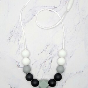 Puede incluir: Un collar con un cord&oacute;n blanco y una variedad de cuentas. Las cuentas son blancas, grises y negras, con una cuenta geom&eacute;trica verde claro en el centro. El collar se muestra sobre una superficie de m&aacute;rmol blanco y gris.