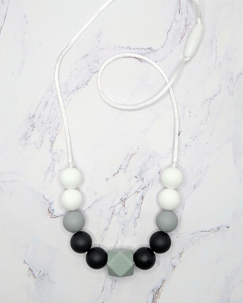 Puede incluir: Un collar con un cord&oacute;n blanco y una variedad de cuentas. Las cuentas son blancas, grises y negras, con una cuenta geom&eacute;trica verde claro en el centro. El collar se muestra sobre una superficie de m&aacute;rmol blanco y gris.