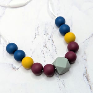 Puede incluir: Un collar de dentici&oacute;n colorido con cuentas redondas azul marino, amarillas y burdeos, y una forma geom&eacute;trica gris. El collar est&aacute; ensartado en un cord&oacute;n blanco.