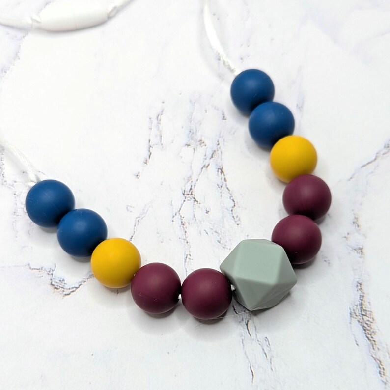 Puede incluir: Un collar de dentici&oacute;n colorido con cuentas redondas azul marino, amarillas y burdeos, y una forma geom&eacute;trica gris. El collar est&aacute; ensartado en un cord&oacute;n blanco.