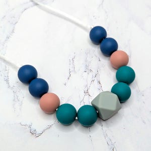 Puede incluir: Un collar con una cadena de cuentas de colores. El collar incluye cuentas redondas azul marino, turquesa y melocot&oacute;n, junto con una cuenta gris de forma geom&eacute;trica. Las cuentas est&aacute;n ensartadas en un cord&oacute;n blanco.