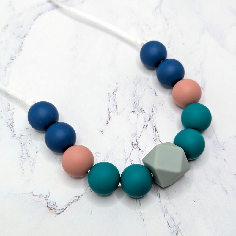 Puede incluir: Un collar con una cadena de cuentas de colores. El collar incluye cuentas redondas azul marino, turquesa y melocot&oacute;n, junto con una cuenta gris de forma geom&eacute;trica. Las cuentas est&aacute;n ensartadas en un cord&oacute;n blanco.