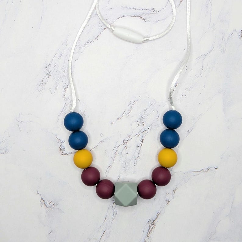 Puede incluir: Un collar colorido con un cord&oacute;n blanco y un cierre de seguridad. Cuenta con cuentas redondas azul marino, amarillas y burdeos, y una cuenta central geom&eacute;trica gris. El collar se exhibe sobre una superficie de m&aacute;rmol blanco.