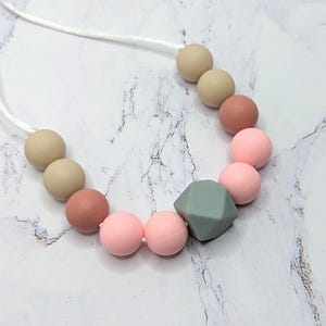 Puede incluir: Un collar con una cadena de cuentas de colores. Las cuentas son en tonos de rosa, beige y una forma geom&eacute;trica verde apagada. El collar est&aacute; ensartado en un cord&oacute;n blanco.