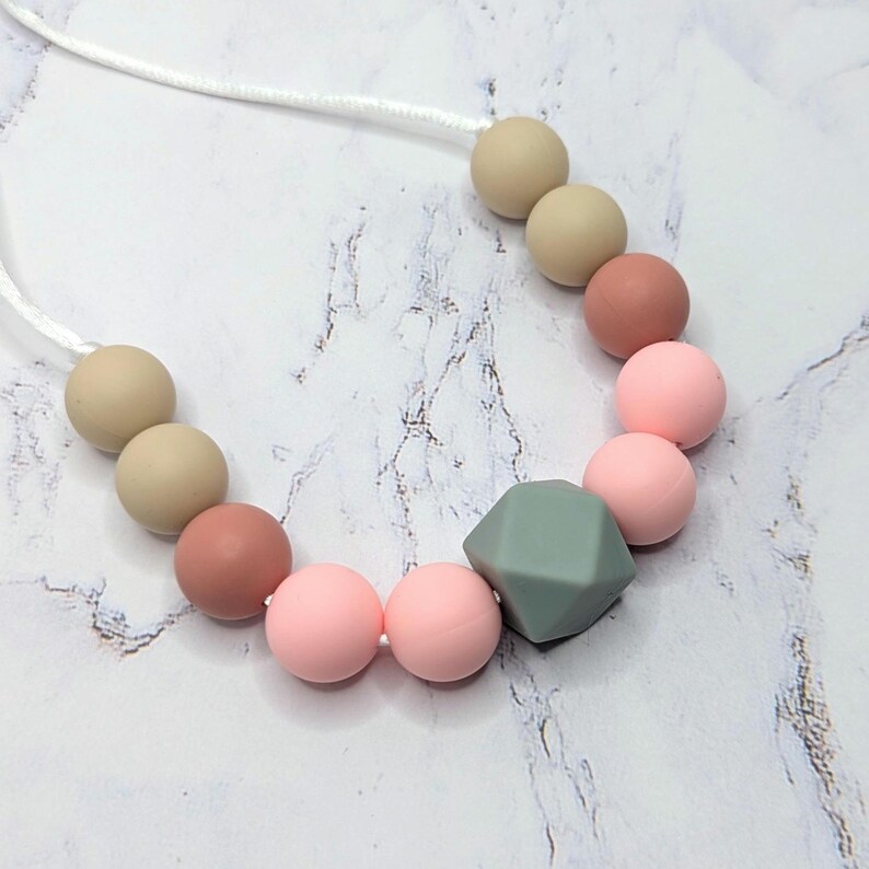 Puede incluir: Un collar con una cadena de cuentas de colores. Las cuentas son en tonos de rosa, beige y una forma geom&eacute;trica verde apagada. El collar est&aacute; ensartado en un cord&oacute;n blanco.
