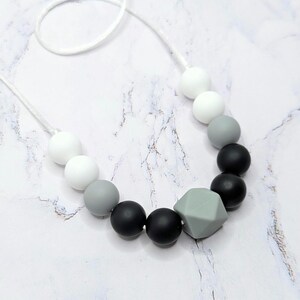 Puede incluir: Un collar con un degradado de cuentas blancas, grises y negras, con un colgante geom&eacute;trico verde claro. Las cuentas redondas van del blanco al gris y al negro. El collar est&aacute; ensartado en un cord&oacute;n blanco.