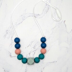 Puede incluir: Un collar para beb&eacute; con un cord&oacute;n blanco y una serie de cuentas de colores. Las cuentas incluyen esferas azul marino, rosa coral y verde azulado, junto con un cubo geom&eacute;trico gris.