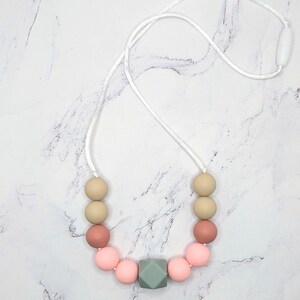 Puede incluir: Un collar con cuentas rosas, beige y verdes. El collar tiene un cord&oacute;n blanco y un cierre blanco. Las cuentas son redondas, con una cuenta geom&eacute;trica en forma de cubo.