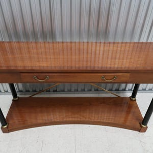 May include: A wooden console table with a warm brown finish. It features a rectangular top, a single drawer with brass-toned handles, and a lower shelf. The legs are black with gold accents. The table is approximately 60 inches long.
