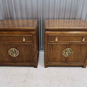 May include: Two matching wooden nightstands with a warm brown finish. Each nightstand features a drawer with a gold-colored handle and a cabinet door with a decorative gold-colored accent. The nightstands have a classic, elegant design.