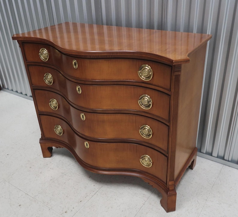 May include: A curved, wooden chest of drawers with four drawers and brass hardware. The chest is a rich brown color and has a traditional style.