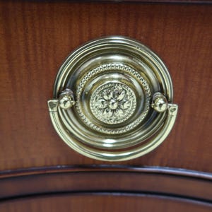 May include: A gold-toned drawer pull with a decorative floral design. The pull is mounted on a wooden surface.
