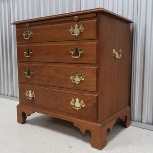 May include: A four-drawer wooden chest of drawers with brass hardware. The chest has a light brown finish and sits on four legs.