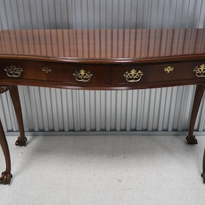May include: A dark brown wooden console table with a curved top and three drawers. The table has ornate legs with carved details and brass hardware.