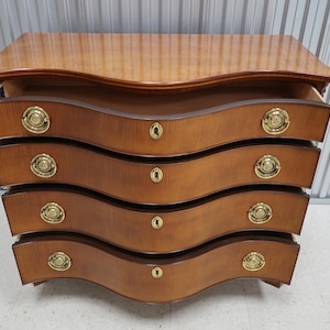 May include: A wooden chest of drawers with four drawers and brass handles. The drawers are curved and the chest has a light brown finish.