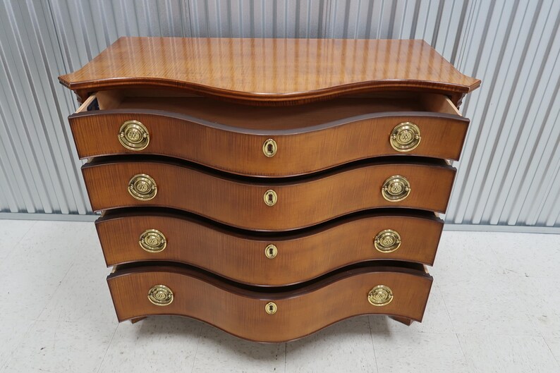 May include: A wooden chest of drawers with four drawers and brass handles. The drawers are curved and the chest has a light brown finish.