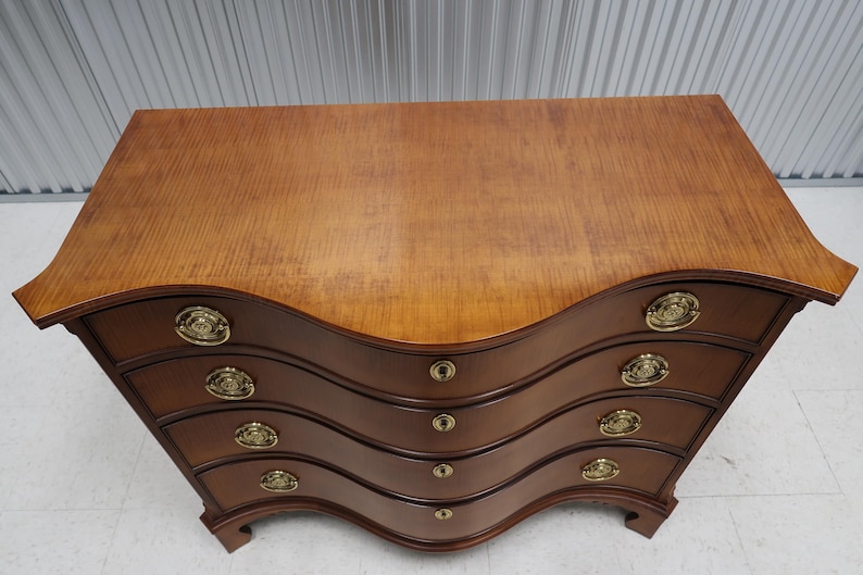 May include: A four-drawer chest of drawers with a curved front and brass hardware. The chest is made of wood with a light brown finish.