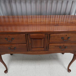 May include: A wooden dresser with a warm brown finish. It features five drawers and a central cabinet door, all with dark metal handles. The dresser has curved legs.