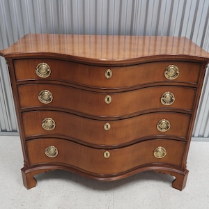 May include: A four-drawer chest of drawers with a curved front and brass hardware. The chest is made of wood and has a light brown finish.