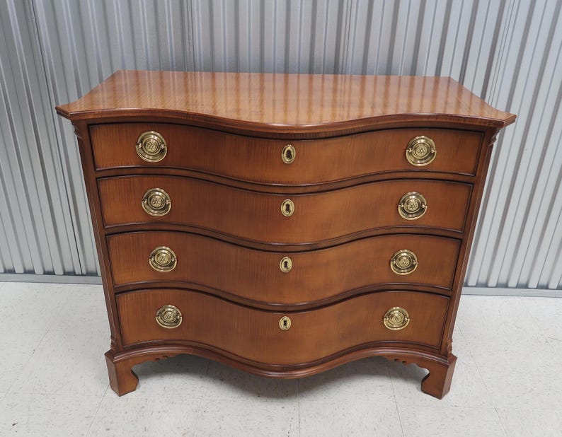 May include: A four-drawer chest of drawers with a curved front and brass hardware. The chest is made of wood and has a light brown finish.