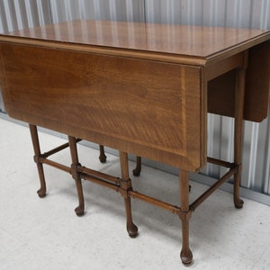 May include: A brown wooden drop leaf table with a rectangular top and a unique, open, rectangular base with four legs. The table is made of wood with a natural finish.